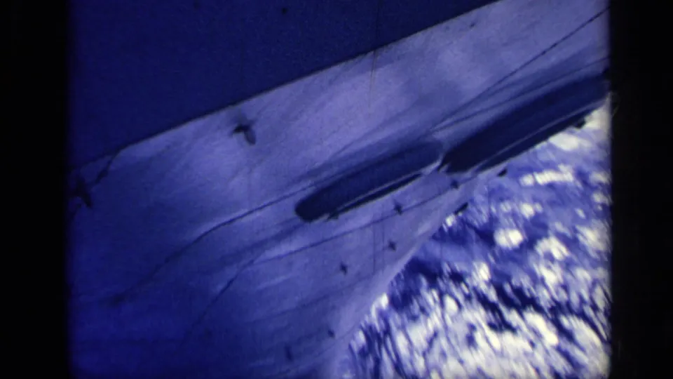 Blue-shifted 1962 Italy home-movie frame of a large vessel's hull pushing past bright, churning water.