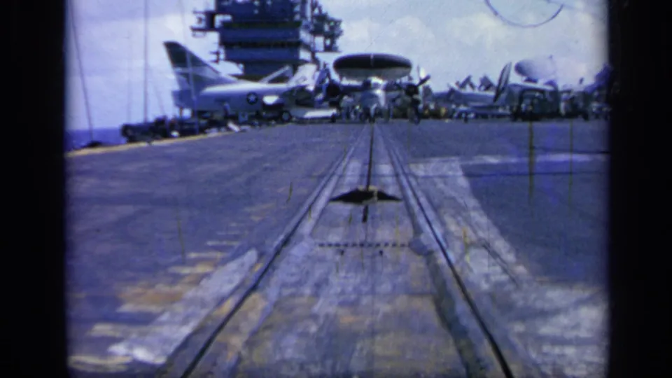 1962 Italy 8mm view along an aircraft carrier deck toward catapult rails, parked military aircraft, and the carrier island.