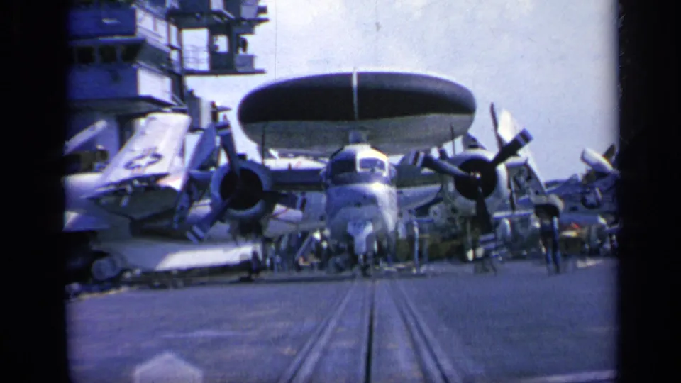 1962 Italy 8mm frame of a radar-equipped propeller aircraft on the carrier deck, with crew figures and nearby aircraft visible.