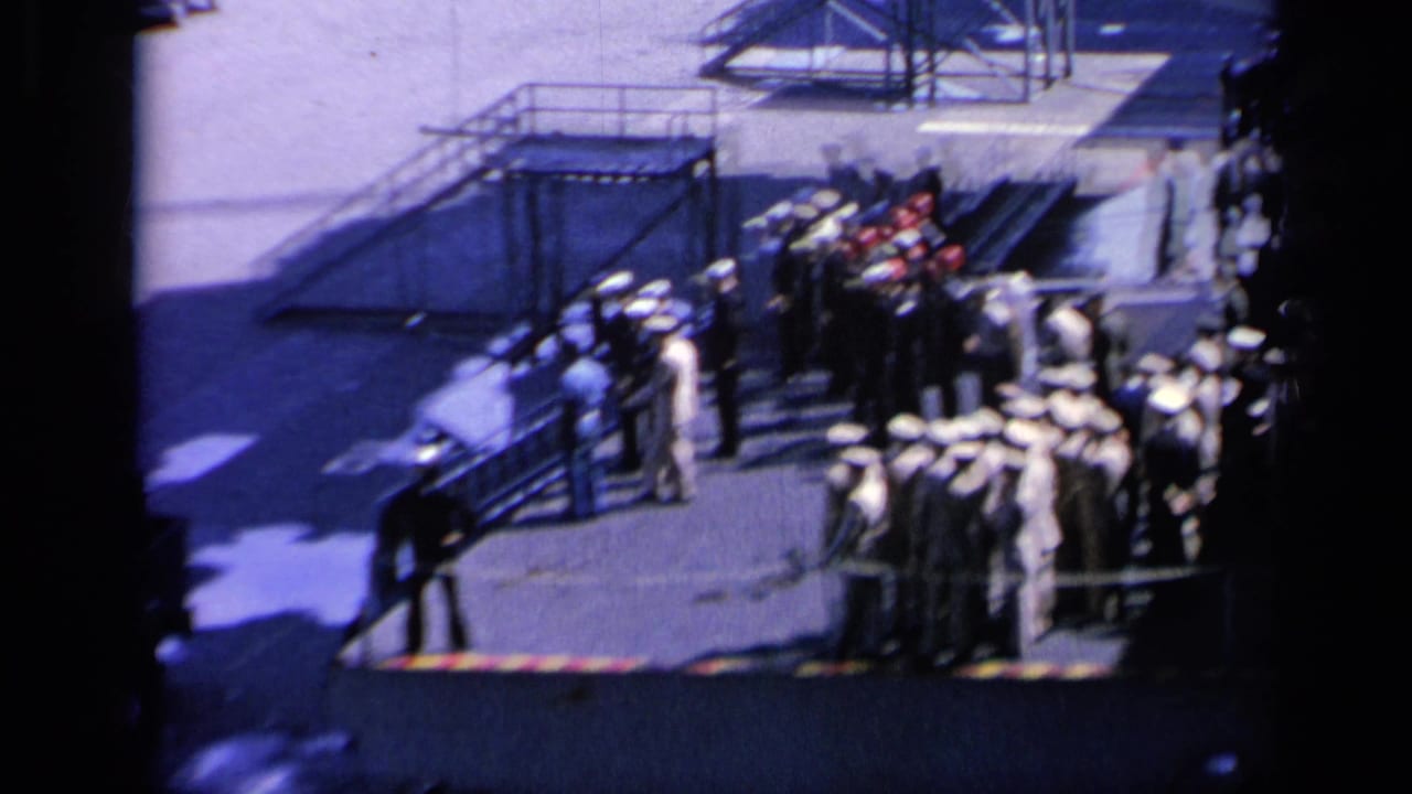 Sailors in white caps line up on a carrier deck near railings and shadowed deck structures.