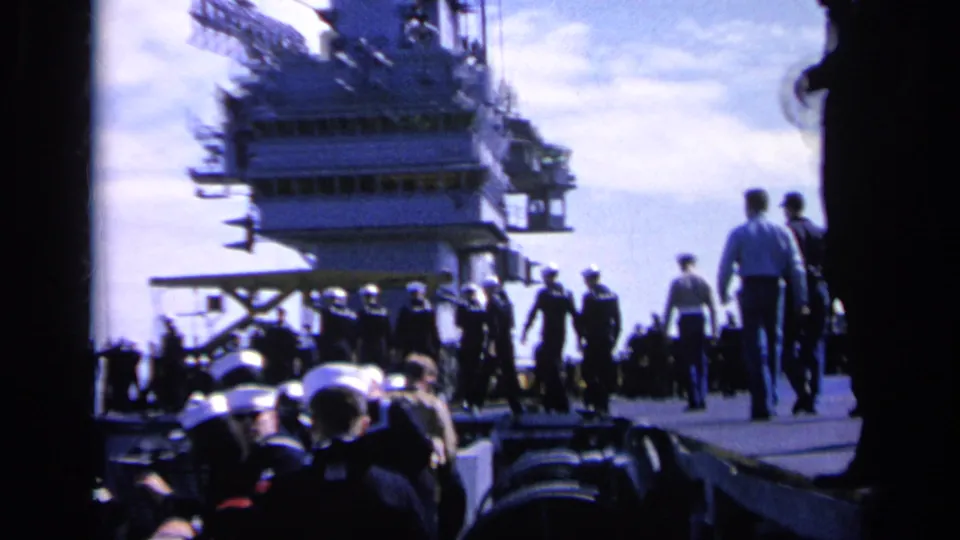 Sailors in dark uniforms and white caps move along an aircraft carrier deck in 1962 Italy footage.