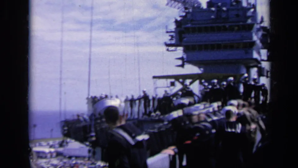 1962 Italy home-movie frame showing sailors on a carrier deck with sea and island structure visible.