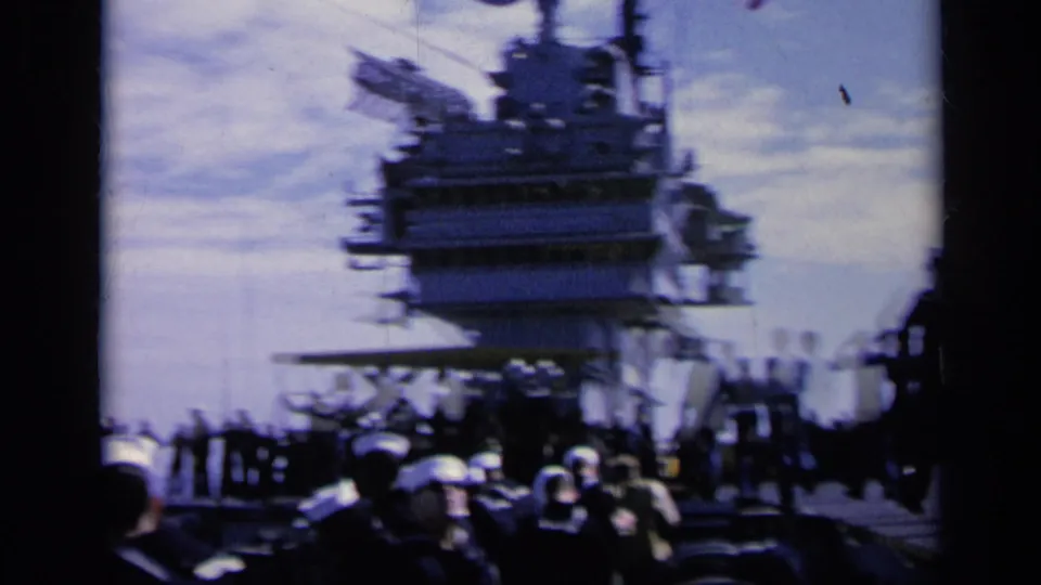 Sailors cluster on an aircraft carrier deck in 1962 Italy footage, with the island superstructure behind them.