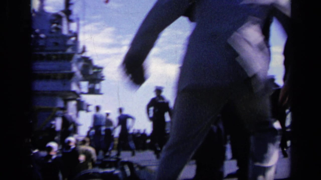Sailors move across a warship deck with ship structure and distant personnel visible.