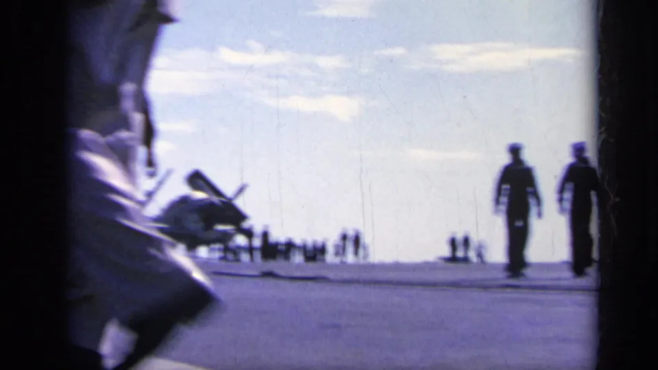 1962 Italy 8mm frame of service members on an open warship deck with a parked aircraft and silhouettes near the horizon.