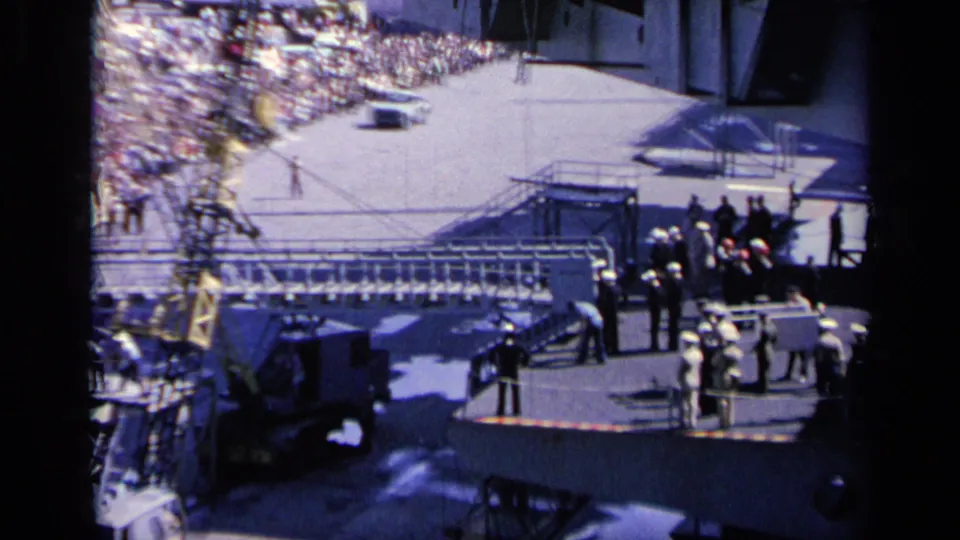 1962 Italy 8mm frame looking down toward a crowded pier, crane rigging, and personnel near the edge of a military vessel.