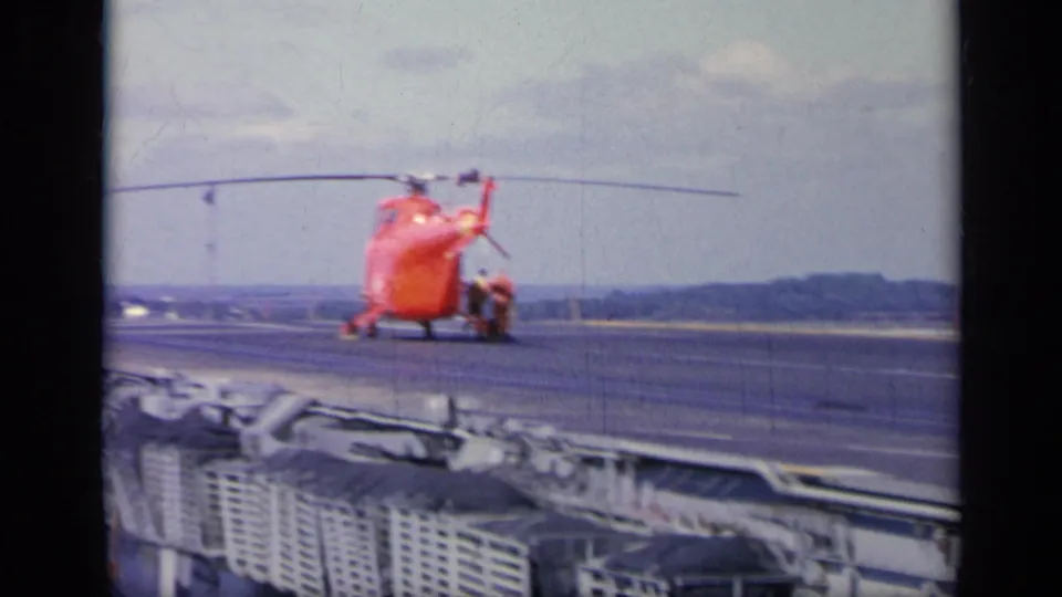1962 Italy frame showing a red helicopter on or near a ship deck, with railings, deck equipment, and water in the background.