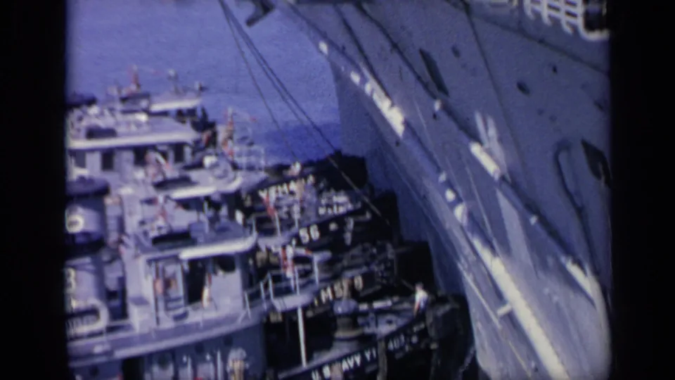 1962 Italy harbor view looking along a large ship toward smaller vessels, deck ladders, rigging, and people working or watching below.