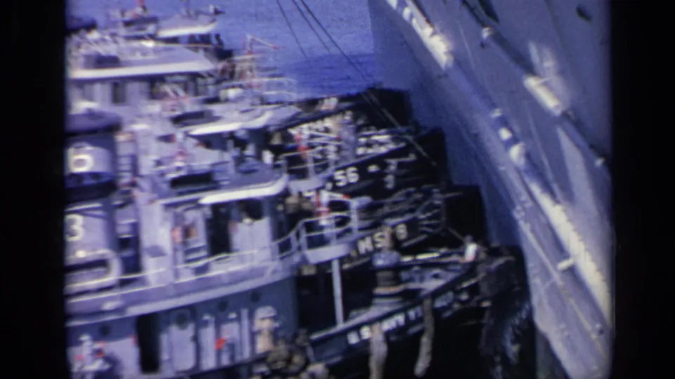 1962 Italy 8mm frame showing gray U.S. Navy tugboats with visible numbers alongside a larger naval vessel in harbor water.