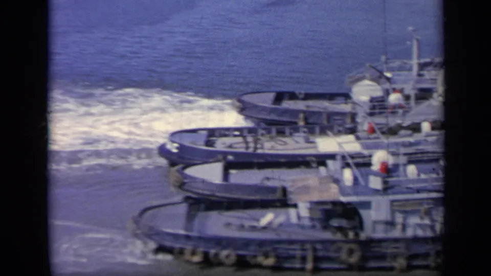 1962 Italy 8mm frame showing a gray Navy tugboat near foamy wake as it maneuvers in harbor water.