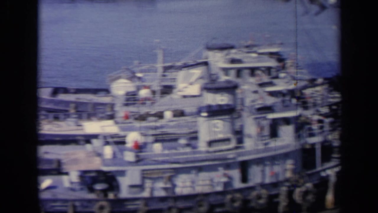 Docked military vessels and numbered tugboats seen from above near harbor water.