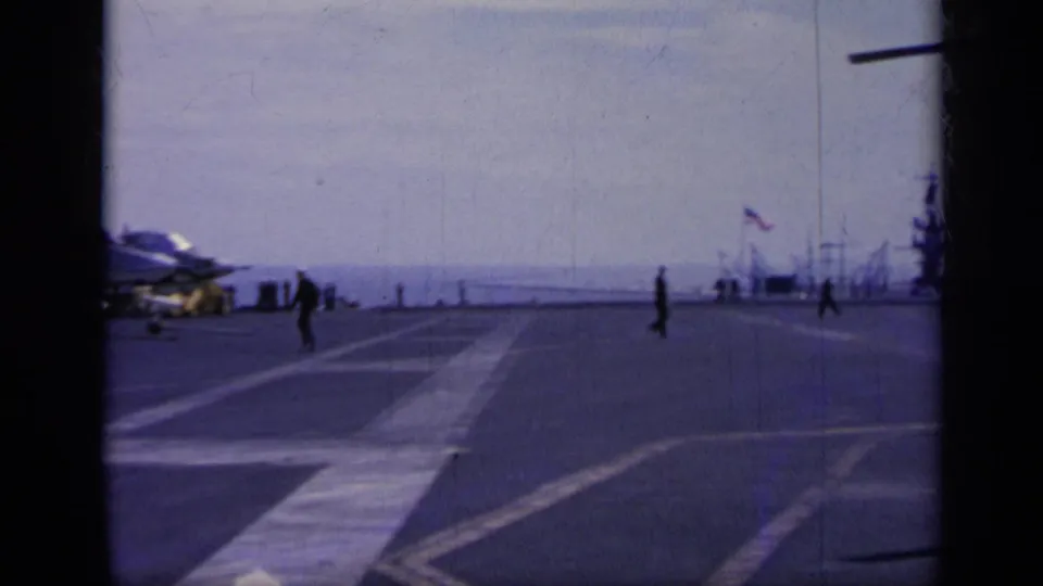 1962 Italy 8mm view across an aircraft carrier flight deck with military personnel walking and aircraft partly visible at the sides.