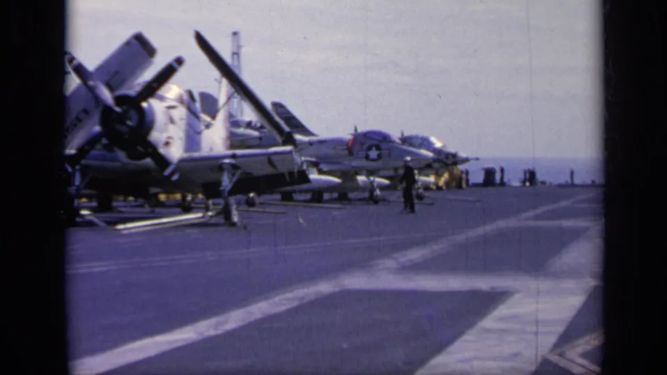 Parked propeller military aircraft sit on a carrier deck in 1962 Italy footage, with personnel walking nearby and the sea beyond.