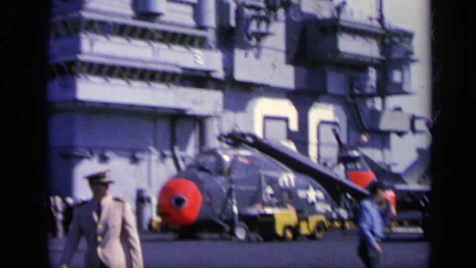 1962 Italy frame of a red-nosed naval aircraft on a carrier deck, with crew and support equipment near the ship island.