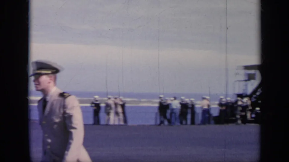 1962 Italy frame showing a uniformed officer in profile, sailors near the deck edge, water, and aircraft equipment nearby.
