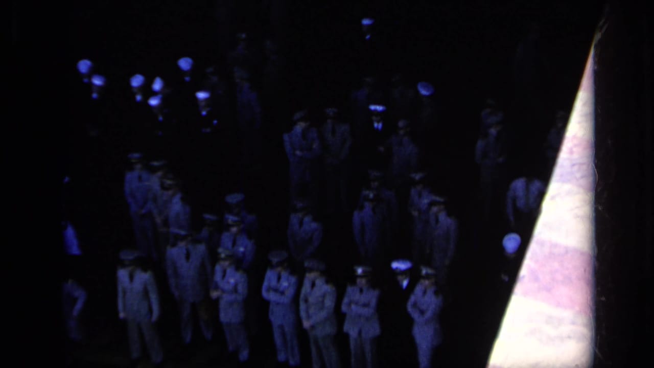 Uniformed Navy personnel standing in a dark ceremony setting with blue lights overhead.