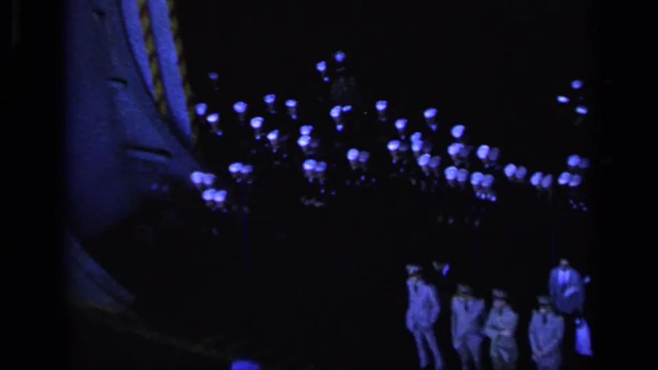 A dim 8mm frame shows formal Navy personnel near a large dark foreground shape and clusters of blue lights.