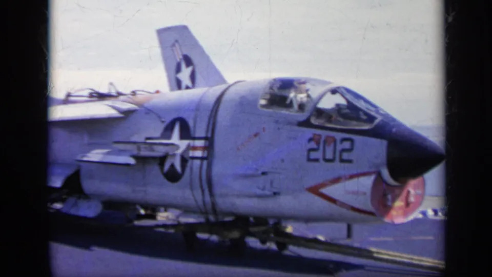 1962 Italy-labeled home-movie frame showing a U.S. Navy jet marked 202 parked on a carrier deck with star-and-bar insignia.