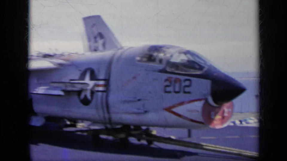 1962 Italy-labeled home-movie frame with a soft, grainy close view of U.S. Navy jet 202 and its aircraft insignia on deck.