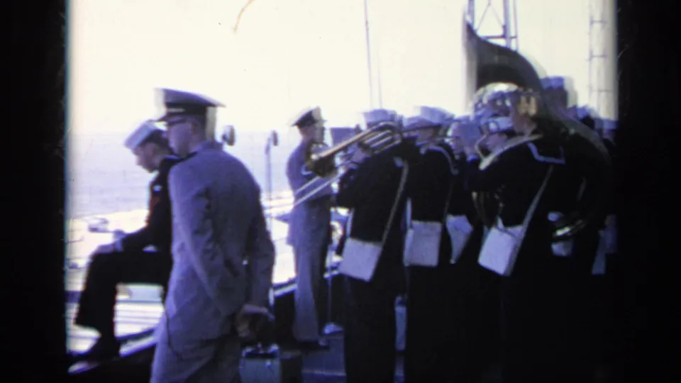 1962 Italy-labeled frame showing uniformed naval musicians and officers on a carrier deck, with trombones and a tuba visible.