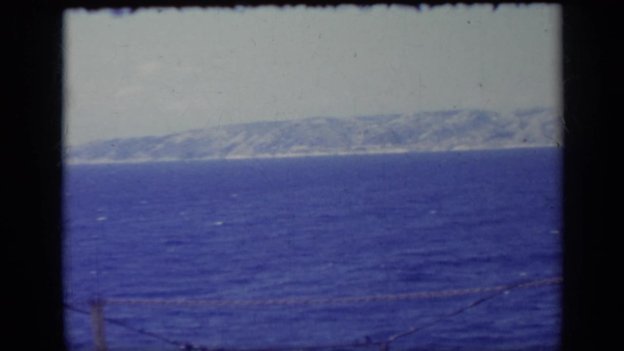 1962 Italy 8mm frame with open sea or lake water, a hazy mountain coastline, and a low foreground cable crossing the view.