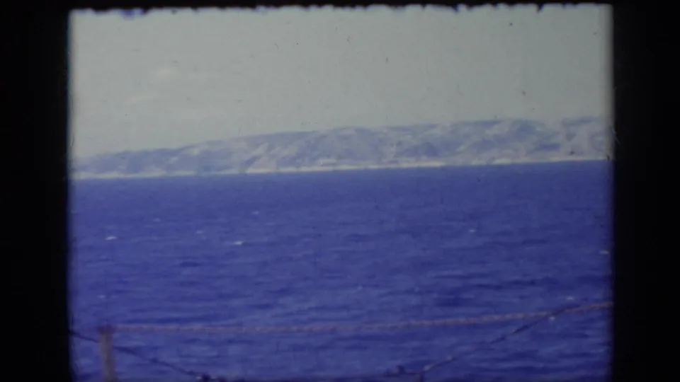 1962 Italy 8mm frame with open sea or lake water, a hazy mountain coastline, and a low foreground cable crossing the view.