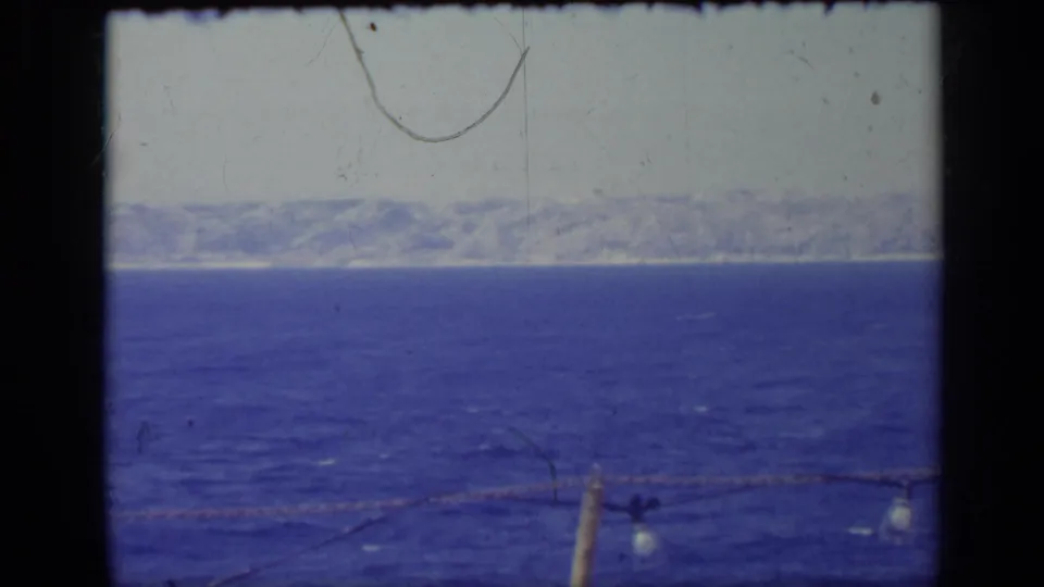 1962 Italy 8mm frame showing a calm blue expanse, distant coast, pale sky, and foreground posts with small bulbs on a rope.