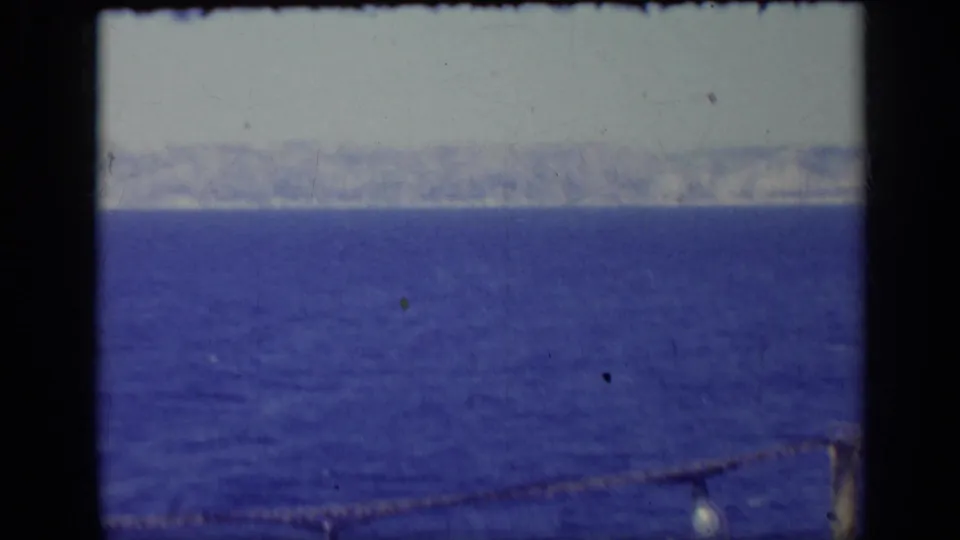 1962 Italy 8mm frame of dark blue water, hazy mountainous coastline, pale sky, and a foreground rope light near the lower edge.