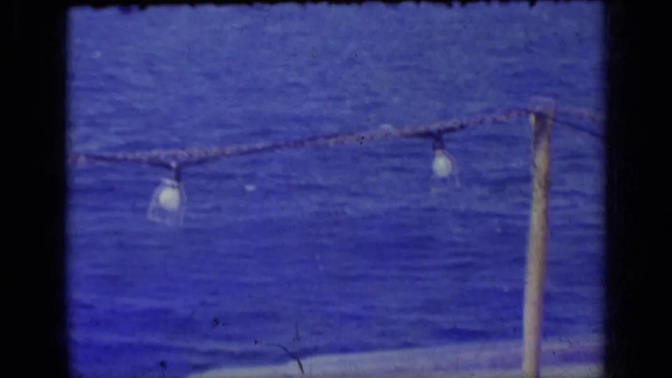1962 Italy home-movie frame showing blue water, a distant mountainous horizon, and hanging rope lights near the camera.