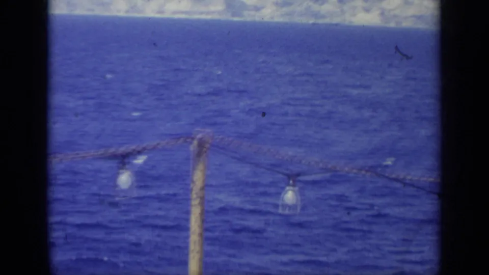 1962 Italy home-movie frame with hanging bulbs strung across a foreground post, open water, and faint land on the horizon.