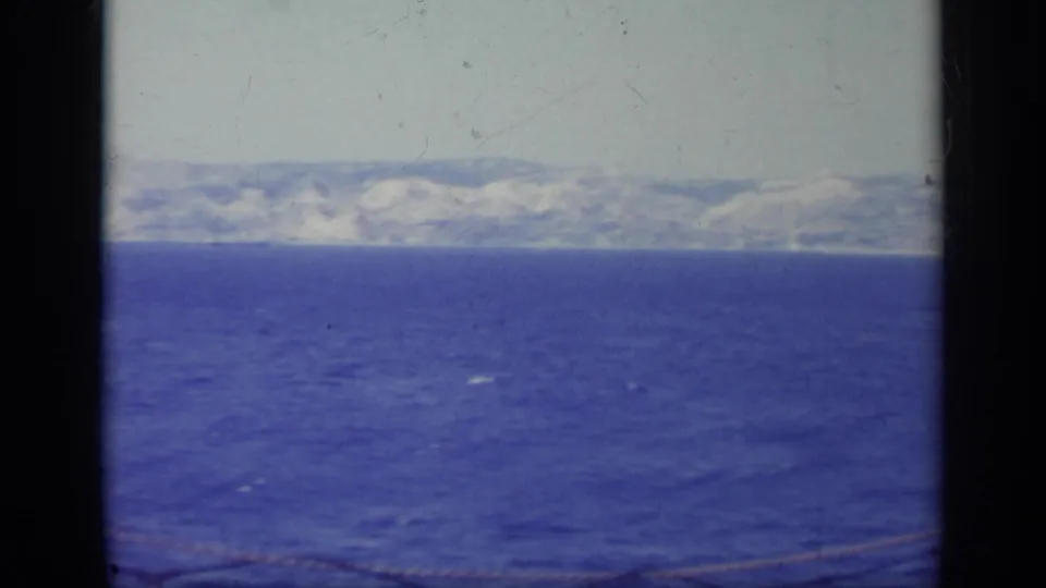 1962 Italy home-movie frame showing a broad blue water surface, pale sky, distant mountains, and partial rope along the lower edge.