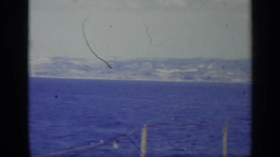 1962 Italy home-movie frame with distant hills across blue water and partial rope lights visible near the bottom of the frame.