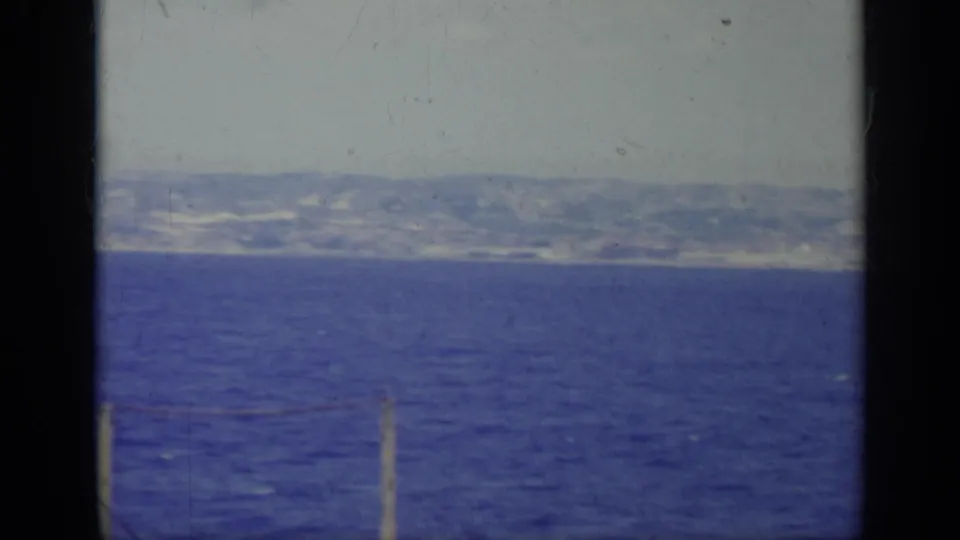 1962 Italy 8mm frame showing blue water, a distant hazy shoreline and mountains, with dark gate edges visible.
