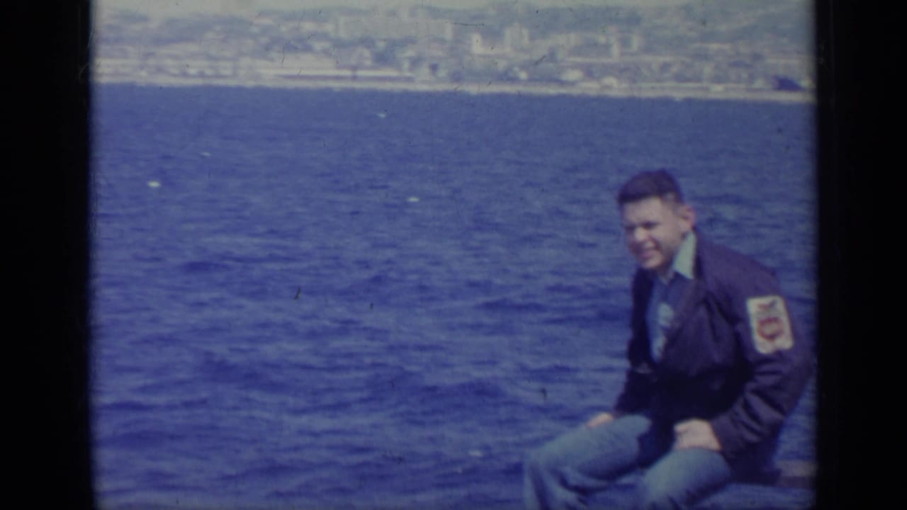 Young man in a dark jacket sitting near blue water with a distant shoreline behind him.