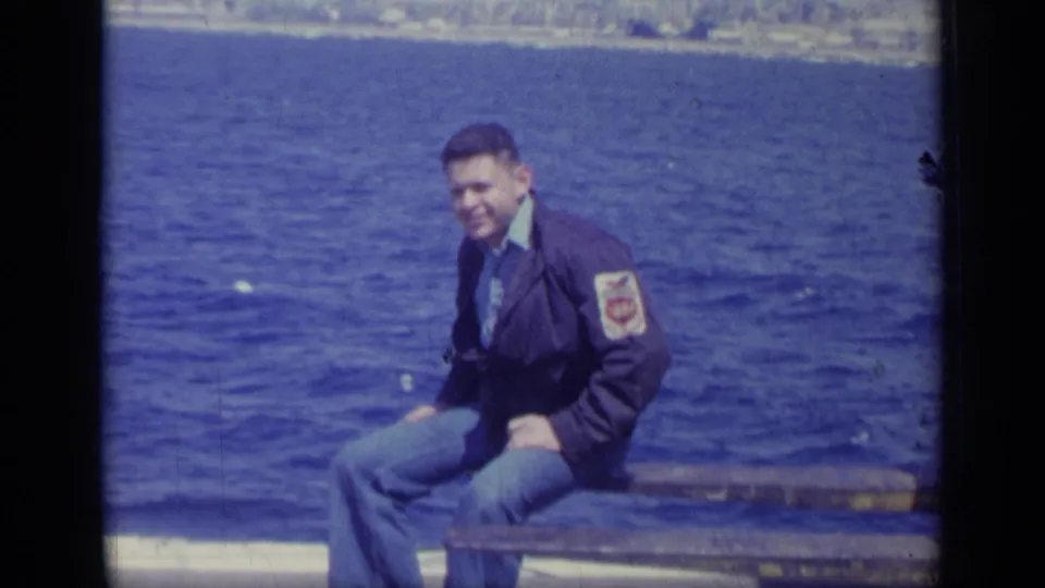 1962 Italy 8mm frame of a smiling young man seated on a ledge by blue water, with a jacket patch visible.