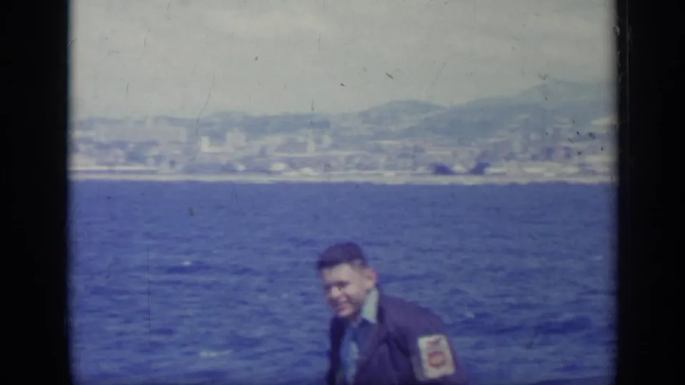 Young man seated near blue water in Italy, with distant shoreline buildings and mountains under a hazy sky.