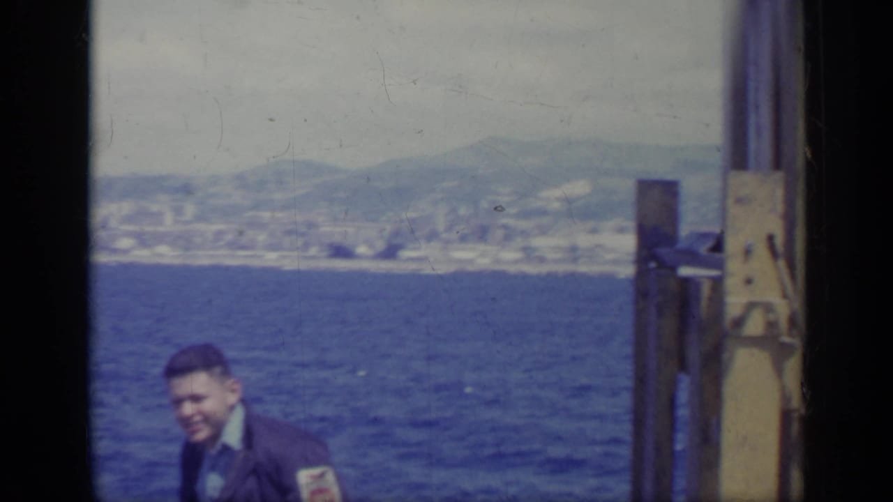 Man in a dark jacket stands on a boat with blue sea and distant coastline behind him.