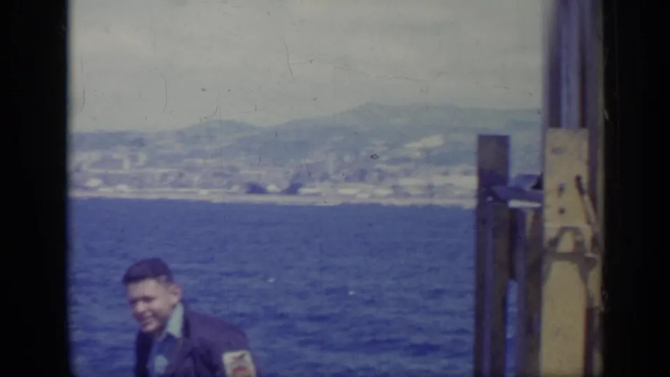 1962 Italy home-movie frame of a man on a boat near a yellow metal structure, with blue water, shoreline, city, and hills beyond.