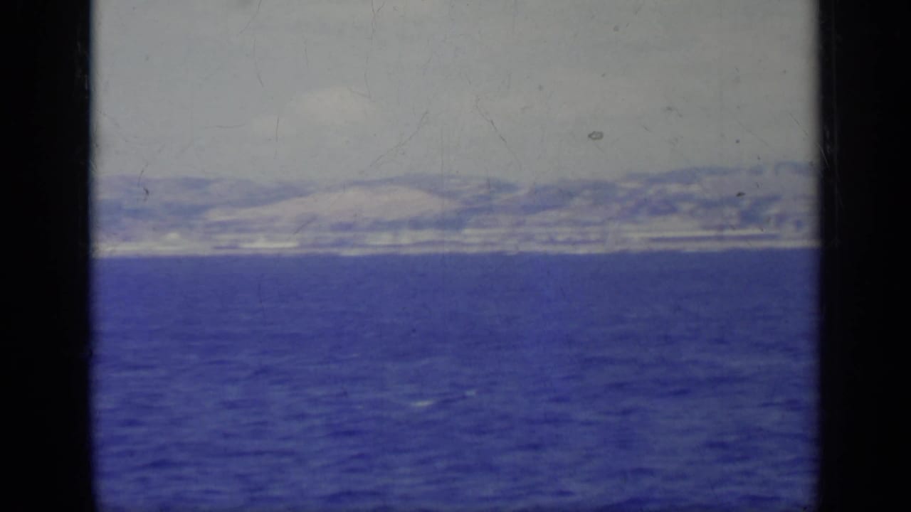 Blue sea and distant shoreline seen past a dark vertical boat frame and yellow deck hardware.