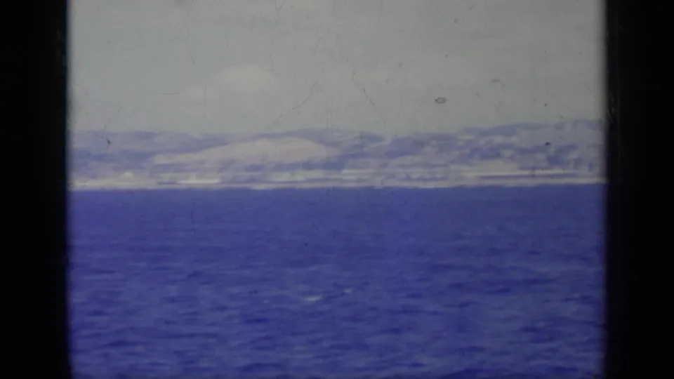1962 Italy home-movie view across blue water toward a pale coastal city and hills, partially blocked by boat framing and metal hardware.