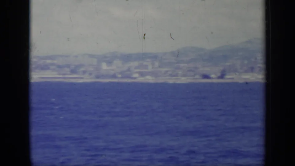 1962 Italy home-movie frame looking from a boat across open water toward a hazy shoreline, city buildings, and low mountains.