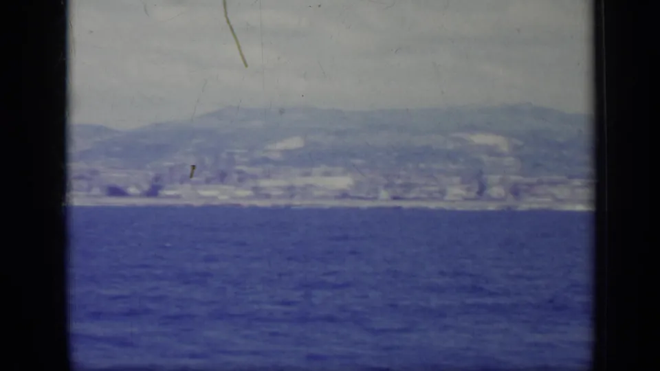 1962 Italy home-movie frame of open blue water leading to a distant coastal city and hills, with soft focus and visible film wear.