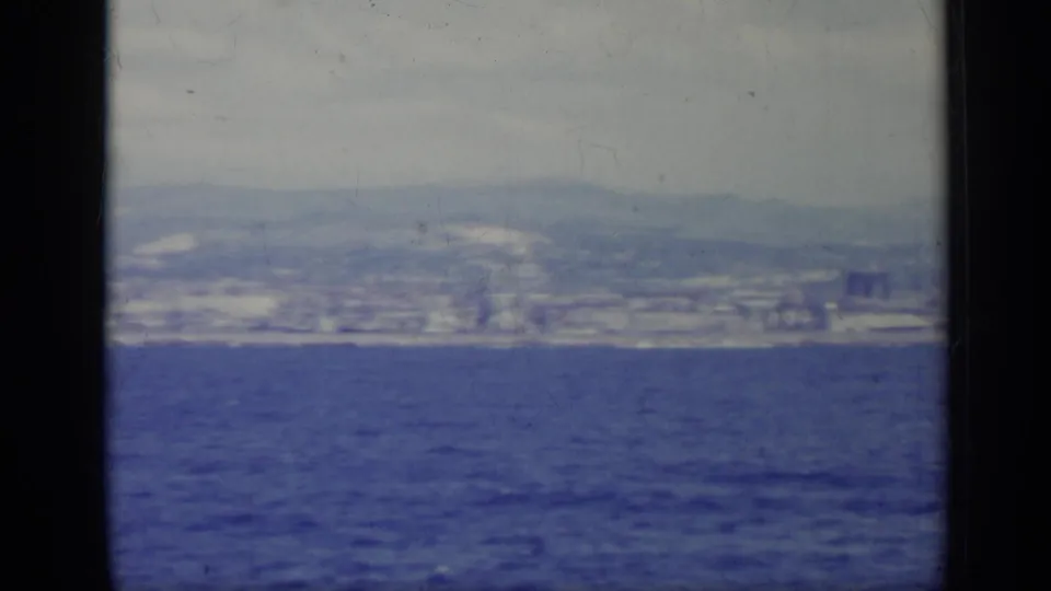 1962 8mm view of an Italian shoreline city seen from offshore, with blue water in the foreground and hazy hills behind.