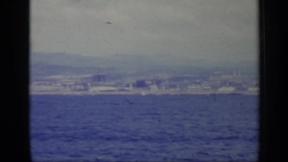 Vintage 8mm frame of a coastal Italian city across the sea, with faint urban forms below hazy mountain slopes.