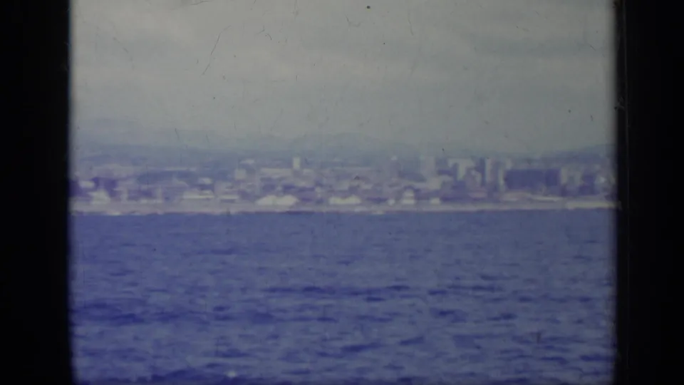 1962 home-movie frame looking from the water toward an Italian coastal city, softened by haze and aged film color.