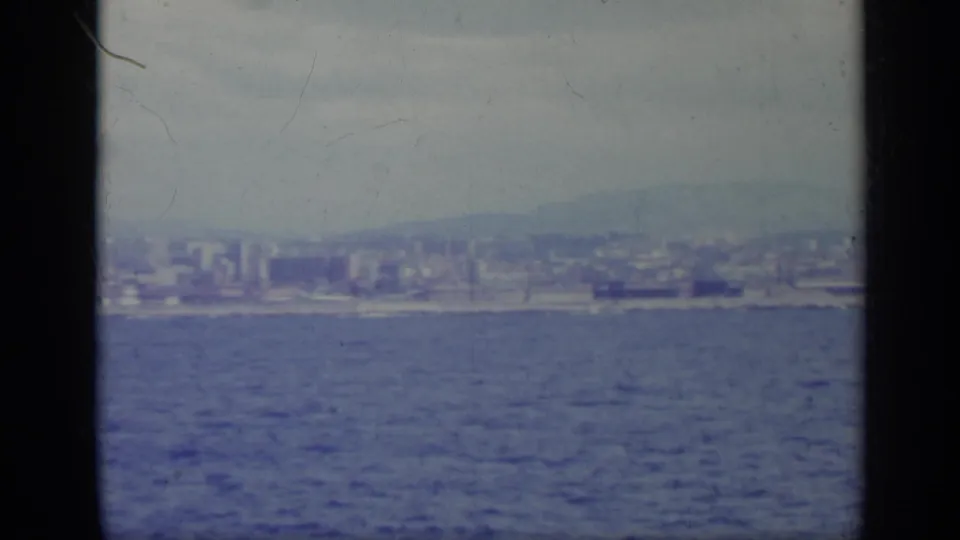 Offshore 8mm view of an Italian waterfront in 1962, showing blue sea, distant buildings, and a muted mountain horizon.
