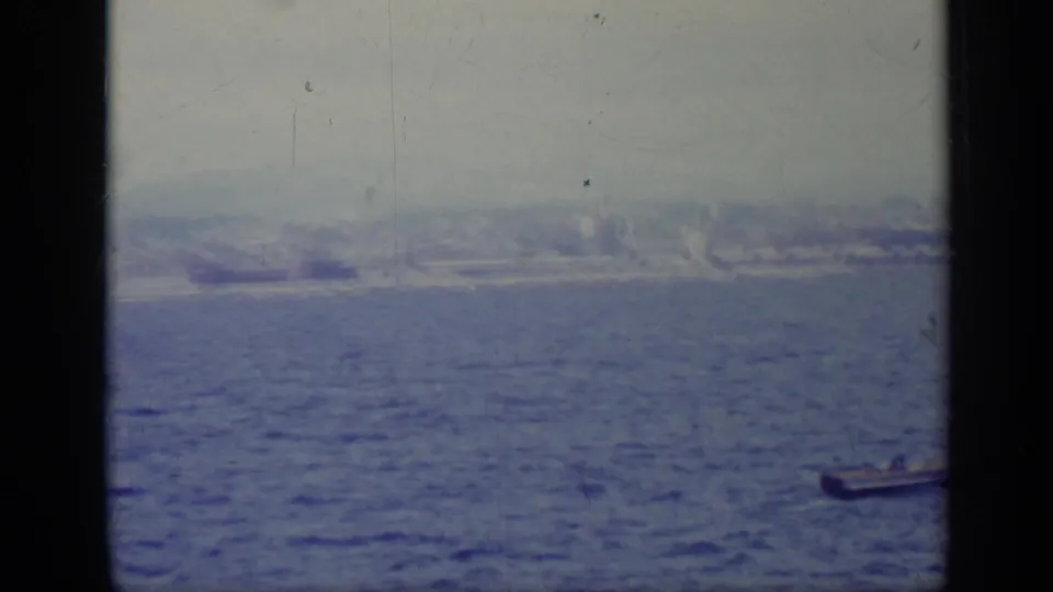 1962 Italy home-movie view across blue water toward a hazy coastal shoreline and distant buildings.