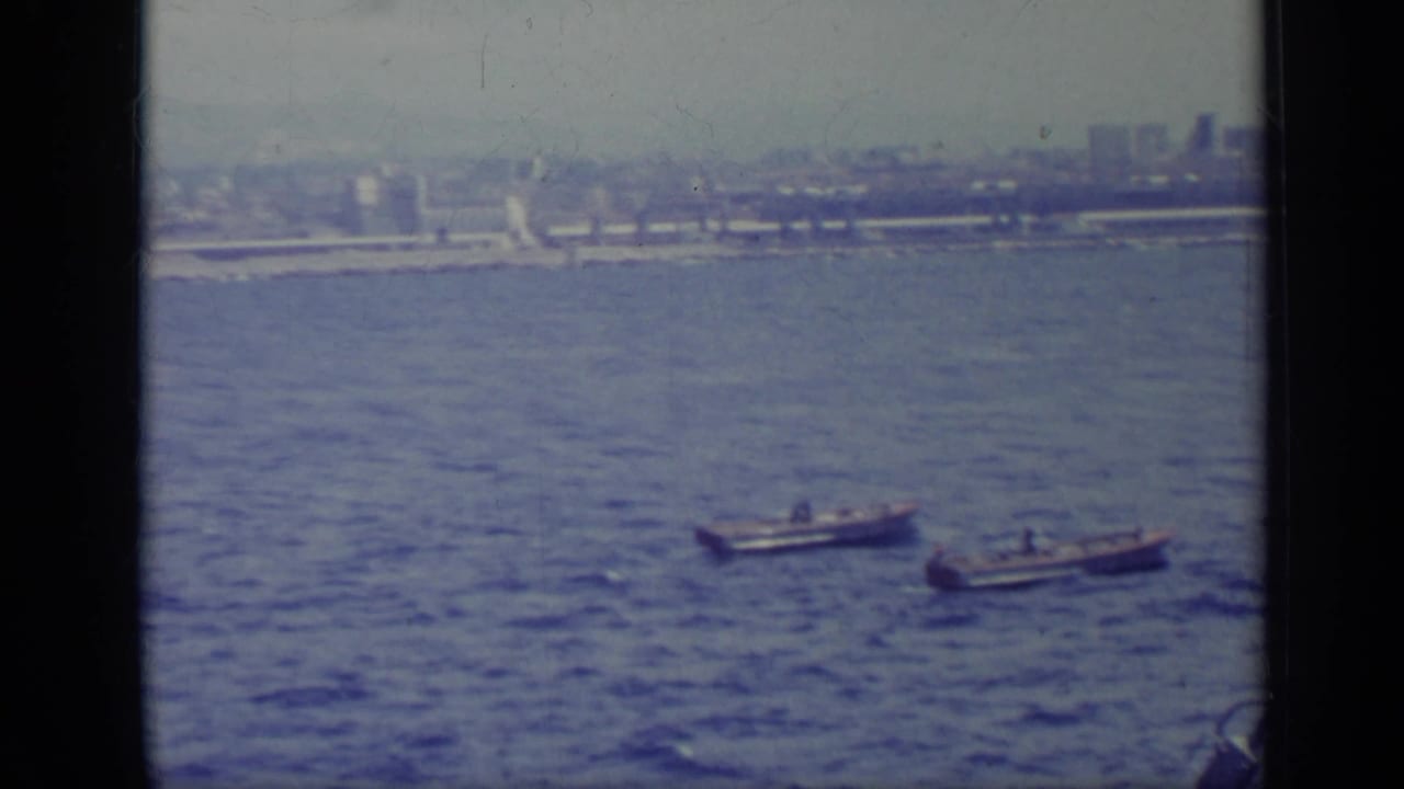 Small boats cross the water in a hazy 1962 Italian coastal view filmed from a ship.