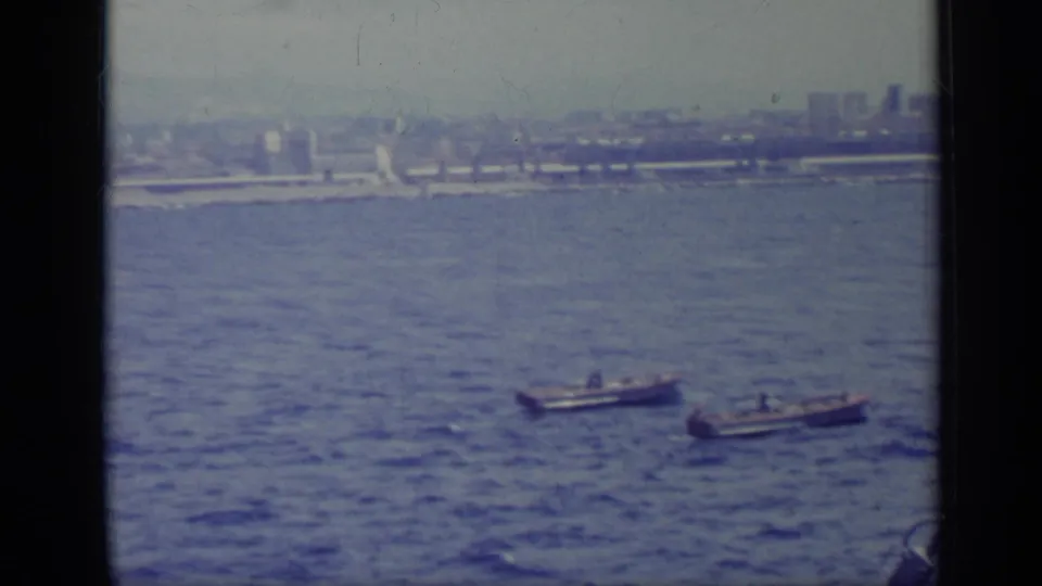 Small boats cross the water in a hazy 1962 Italian coastal view filmed from a ship.