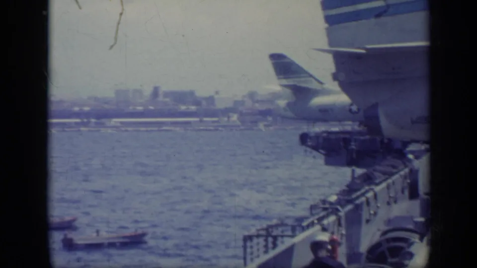 Shipboard view in 1962 Italy showing an airplane tail, a crew member in a white hat, water, boats, and a hazy coast.